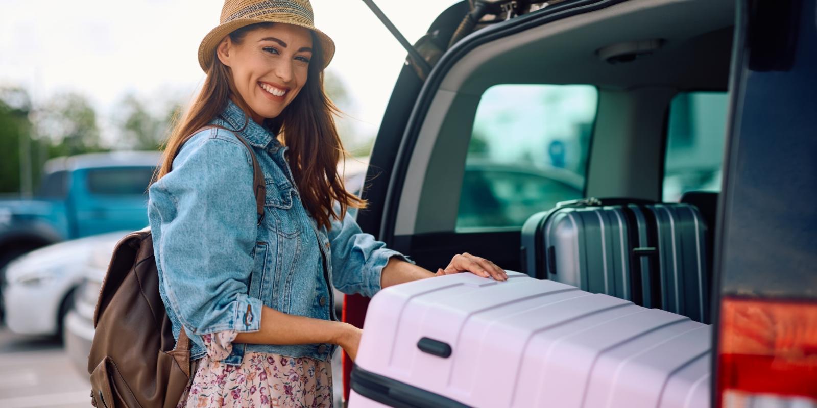 Donna sorridente carica valigia nel bagagliaio di un'auto, pronta per il viaggio.