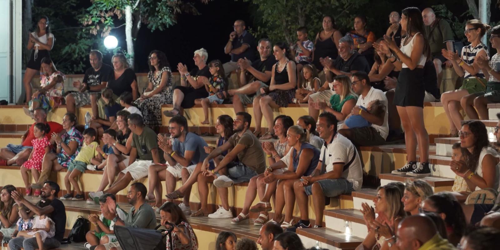Pubblico seduto all'aperto applaude durante uno spettacolo serale.