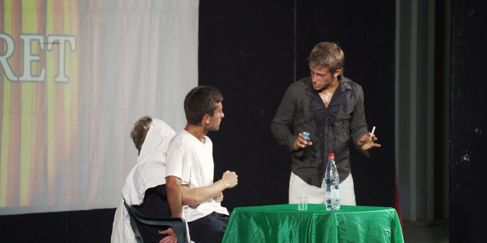 Scena teatrale con tre attori, uno coperto da un lenzuolo bianco.