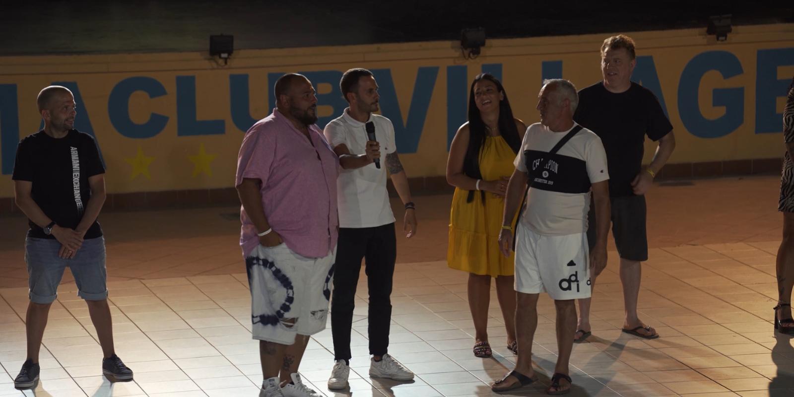 Gruppo di persone su un palco, uno tiene un microfono, sfondo con scritta 'VILLAGE'.