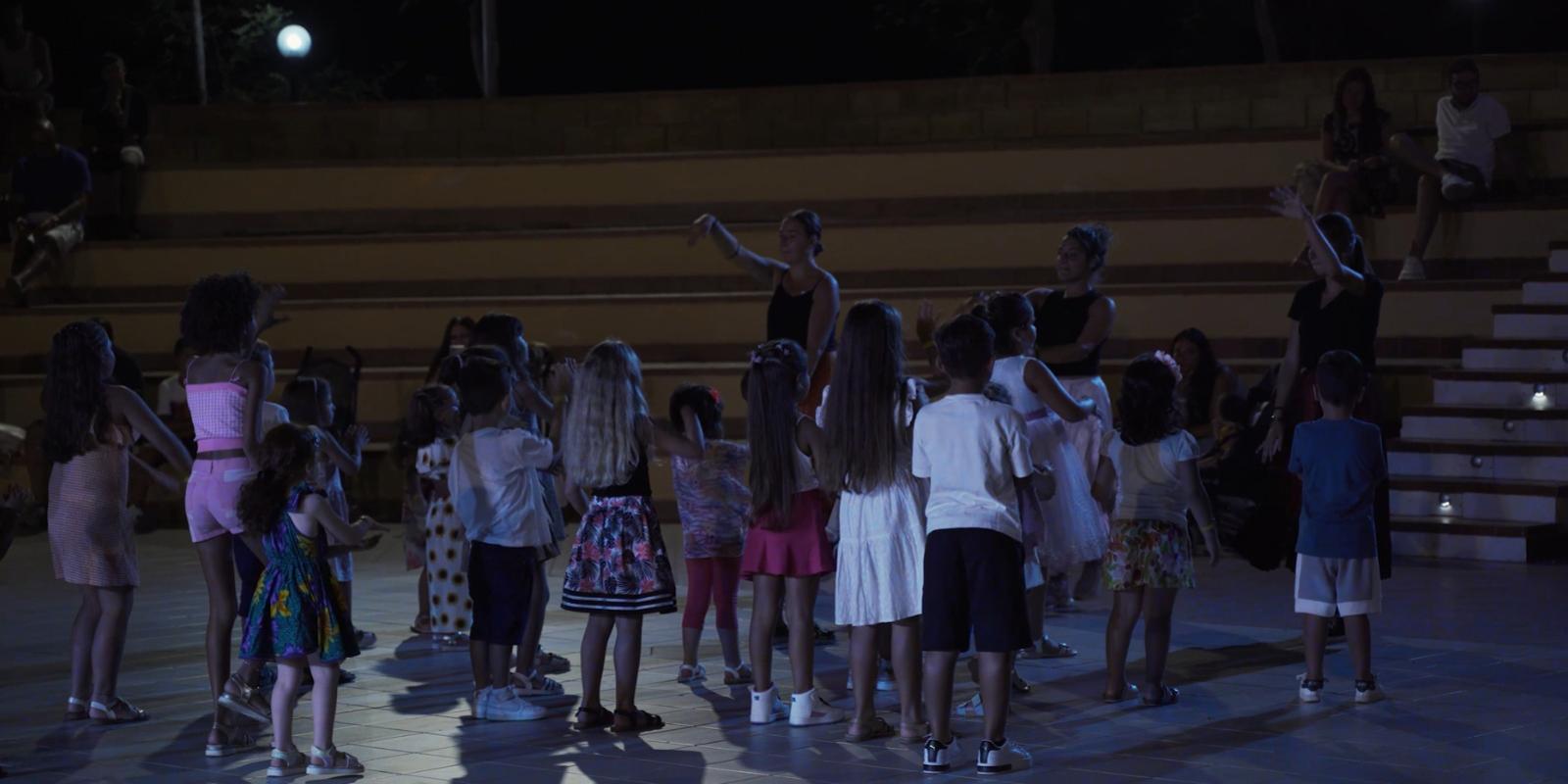 Bambini ballano insieme in un anfiteatro all'aperto di notte, guidati da un istruttore.