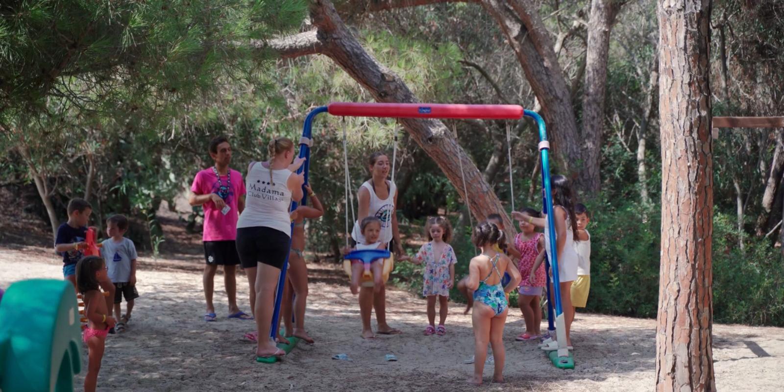 Bambini giocano sull'altalena nel parco, supervisionati da adulti, circondati da alberi.