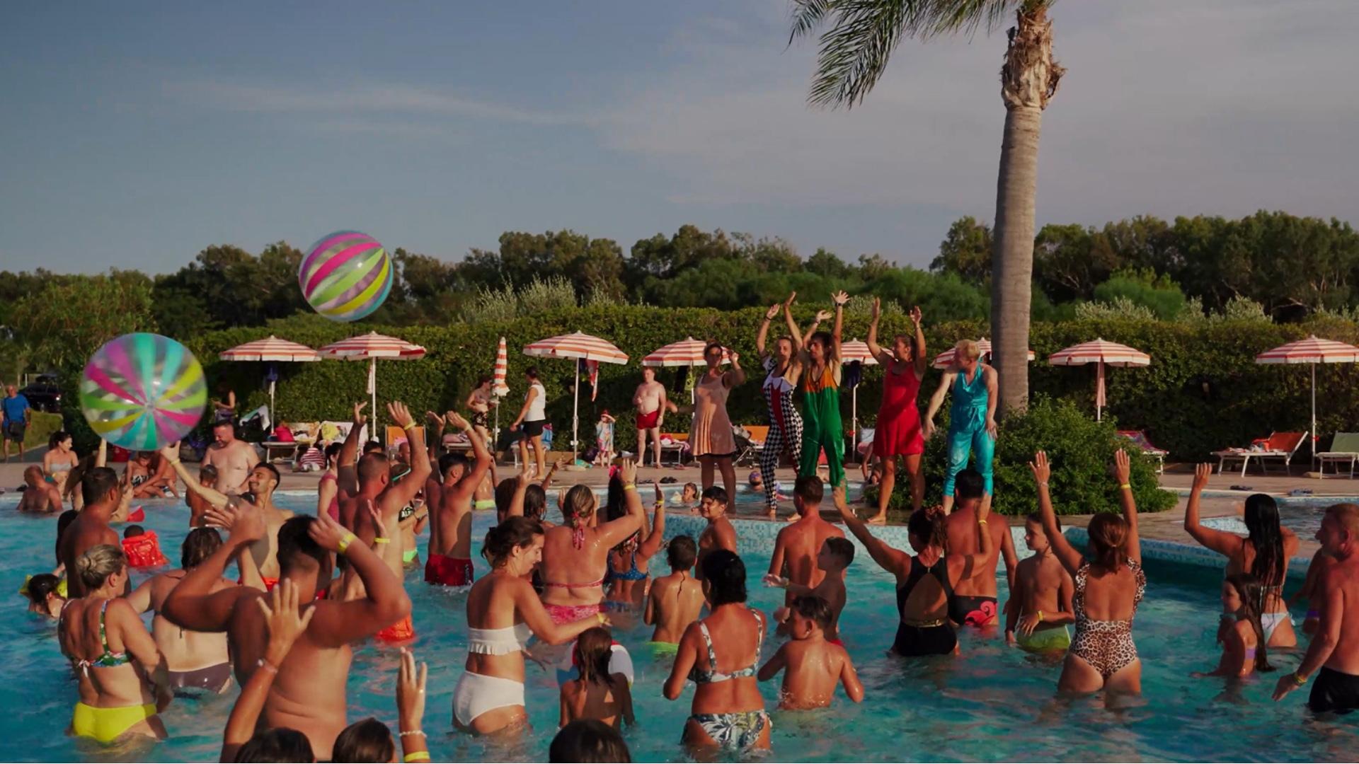 Festa in piscina con animatori e palloni colorati, persone che si divertono nell'acqua.