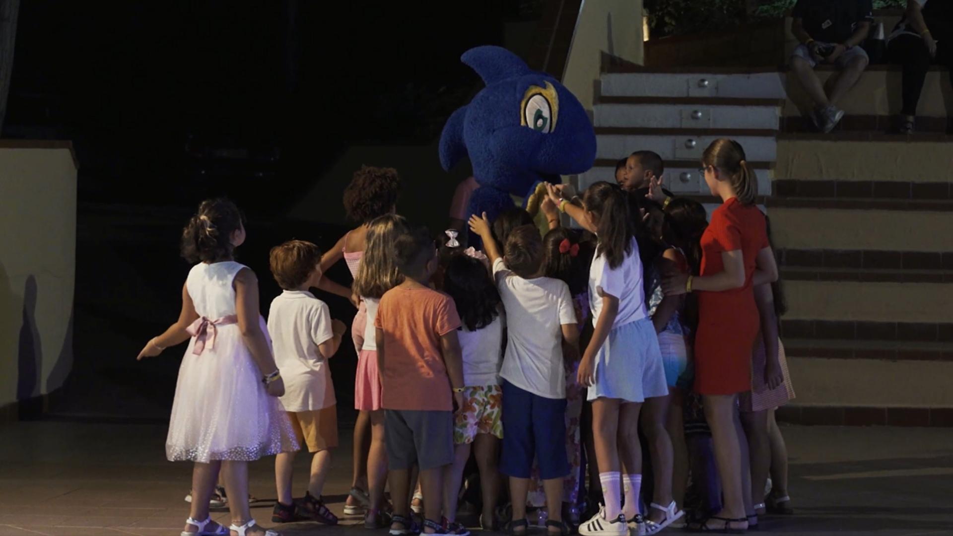 Gruppo di bambini attorno a una mascotte blu durante un evento serale.