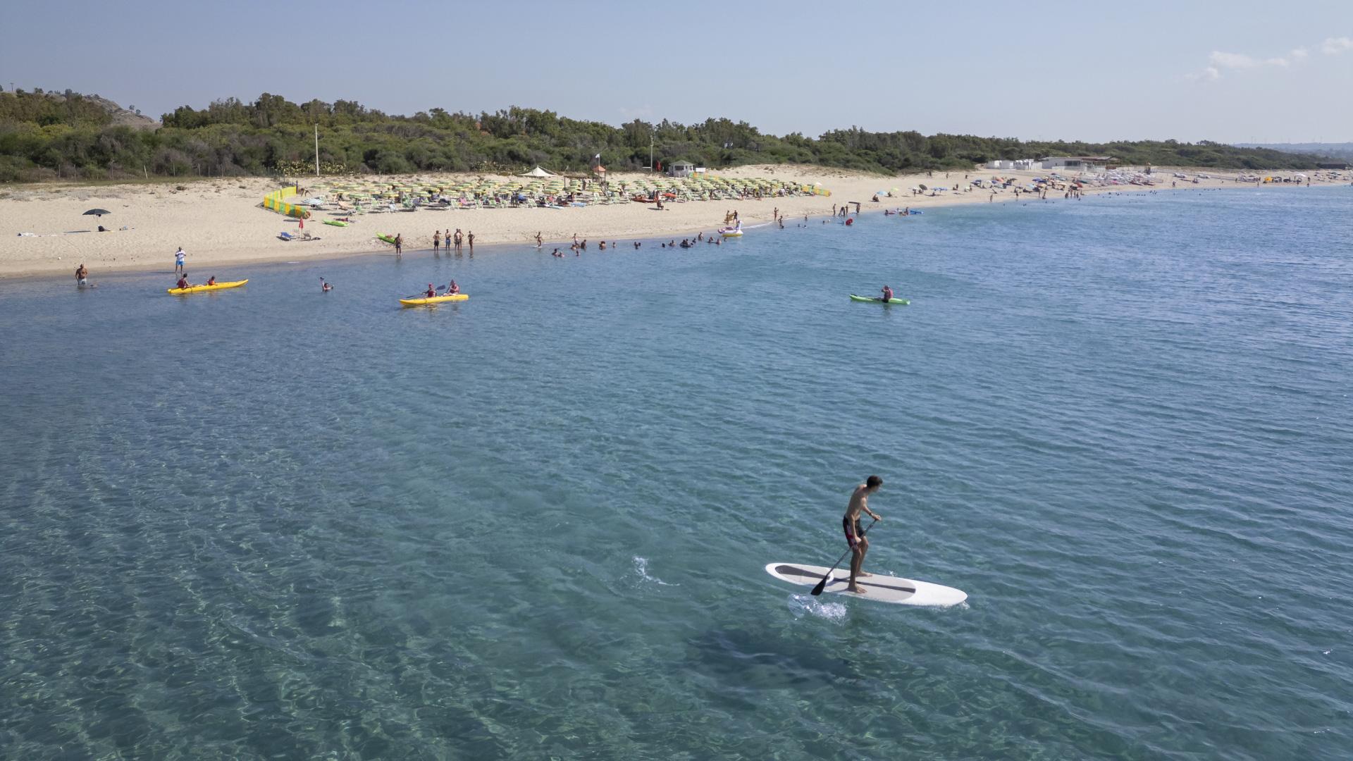 Spiaggia affollata con persone che fanno kayak e paddleboard in mare limpido.