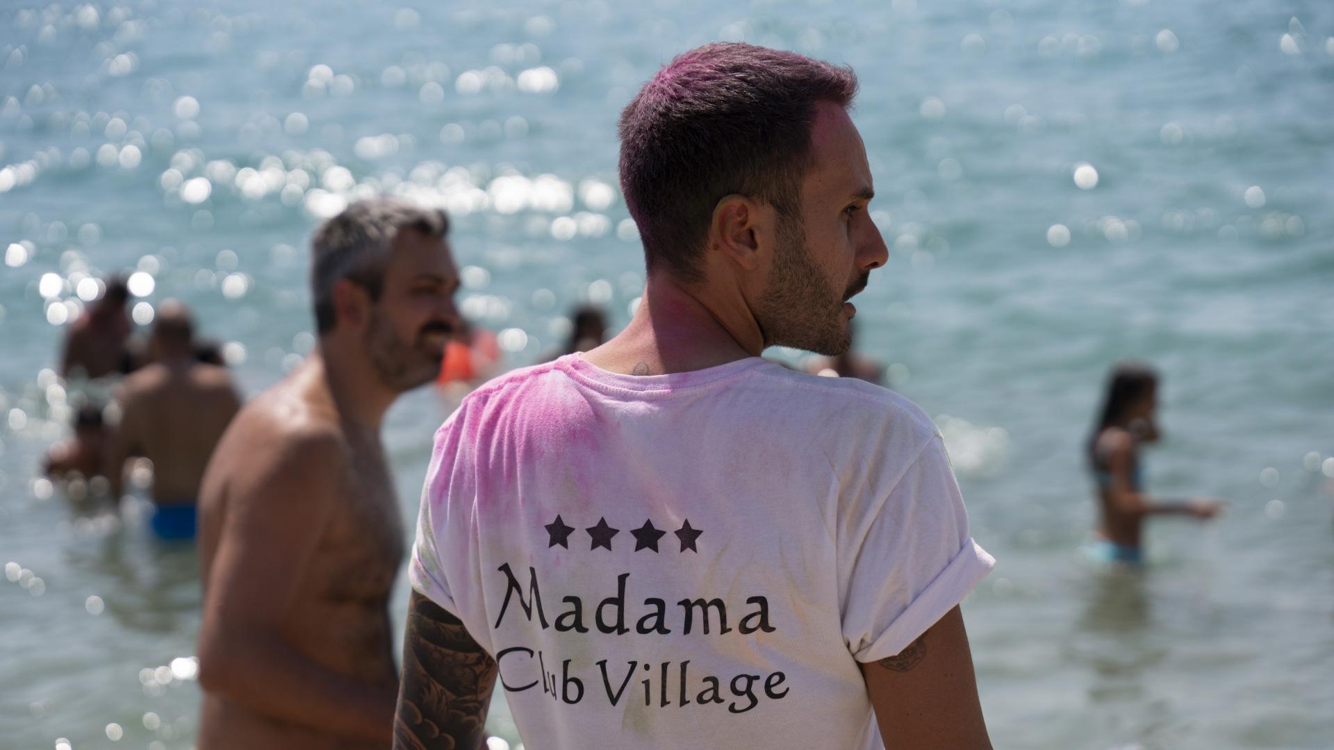 Uomo con maglietta 'Madama Club Village' al mare, circondato da bagnanti.