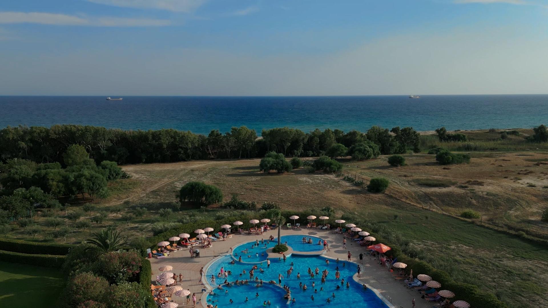 Piscina affollata vicino al mare, circondata da ombrelloni e vegetazione.