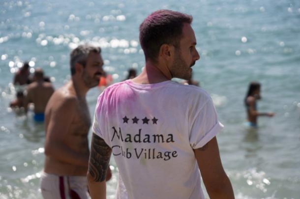 Uomo al mare con maglietta 'Madama Club Village', circondato da bagnanti.