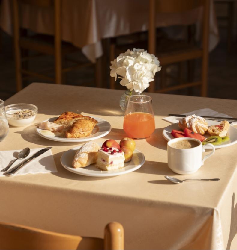 Colazione con dolci, frutta, succo e caffè su tavolo apparecchiato elegantemente.