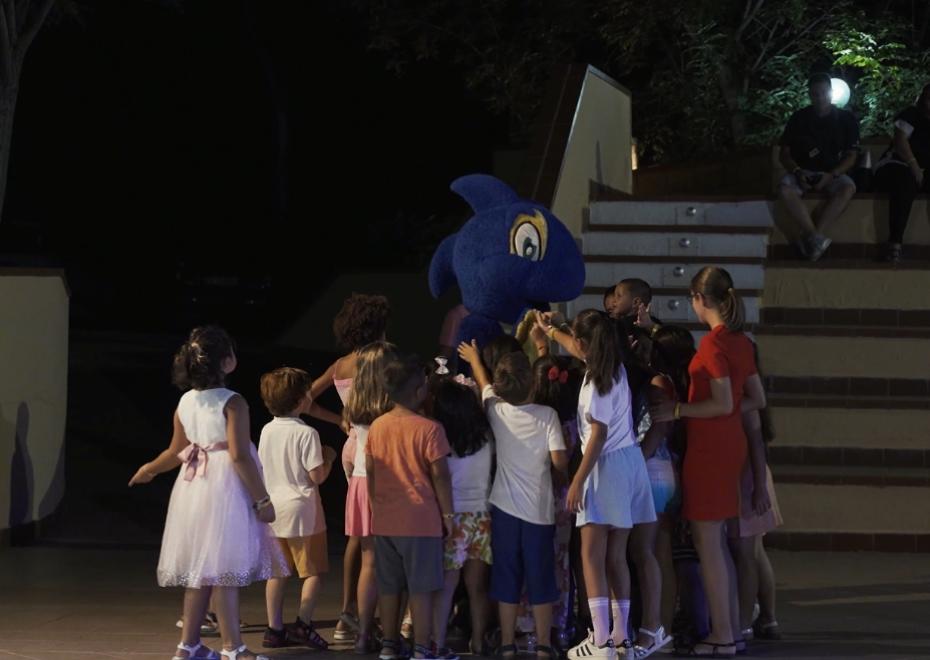 Bambini circondano una mascotte blu durante un evento serale all'aperto.
