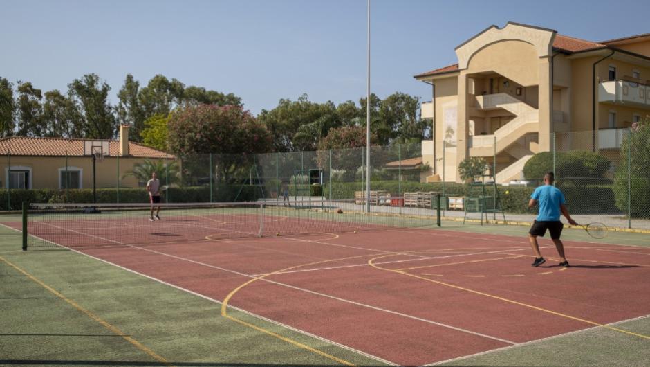 Due persone giocano a tennis su un campo all'aperto, vicino a edifici residenziali.